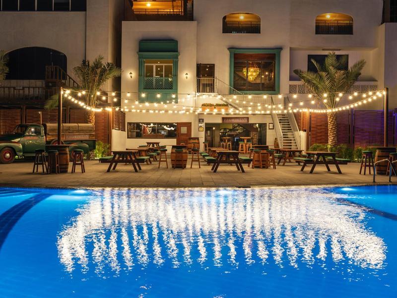 Beleuchtetes Hotel mit Pool, Stühlen und Tischen, Lichterkette spiegelt sich im Wasser wider.