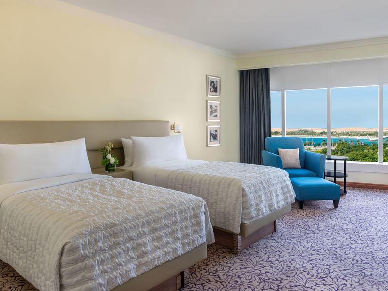 Hotelzimmer mit zwei Einzelbetten, blauem Sessel und großer Fensterfront mit Blick aufs Meer.