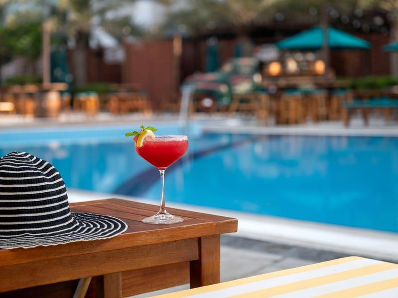 Cocktailglas und gestreifter Sonnenhut auf einem Holztisch neben einem Pool