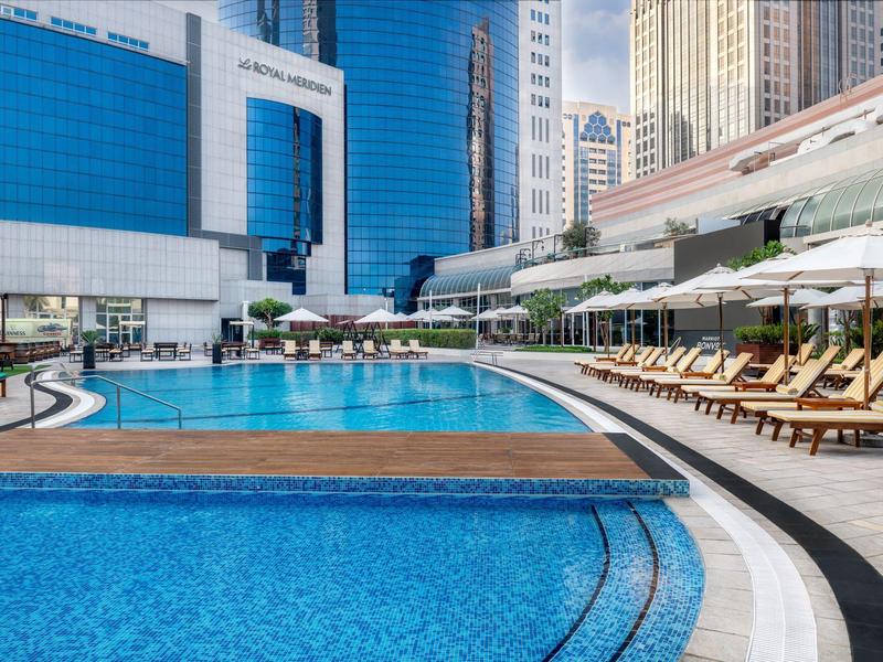 Großer blauer Pool mit Liegestühlen und Sonnenschirmen, umgeben von modernen Hochhäusern.