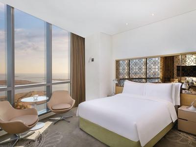 Habitación de hotel moderna con ventana grande, cama cómoda y dos sillones con vista al mar.