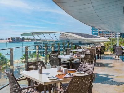Offene Terrasse mit Tischen und Stühlen am Hotel mit Blick aufs Meer und modernen Gebäuden.