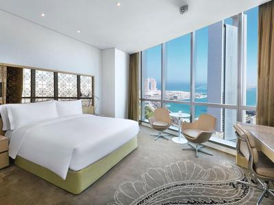Helles Hotelzimmer mit großem Bett, Stühlen am Fenster und Blick auf Meer und Stadt.