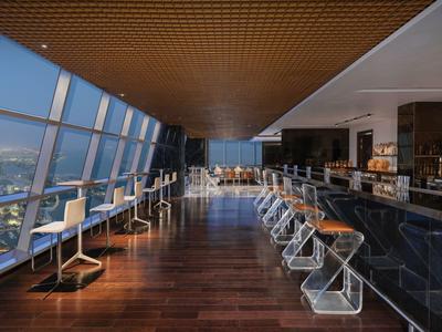 Área moderna de bar de hotel con ventanas altas y vista a la ciudad durante el día.