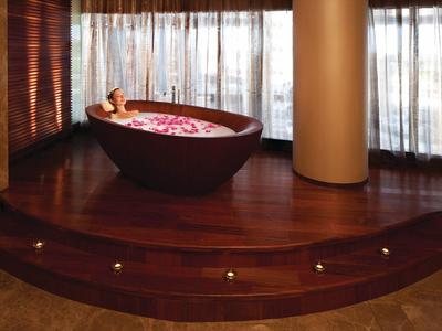 Una persona se relaja en una bañera independiente con pétalos en una plataforma de madera elevada frente a ventanas de piso a techo.