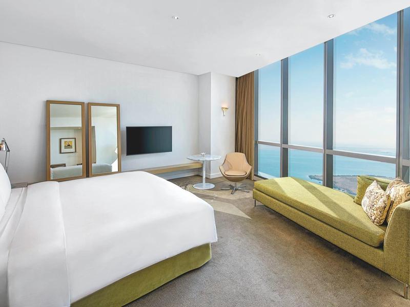 Habitación de hotel moderna con cama grande, sofá, silla y ventanas panorámicas con vista al mar.