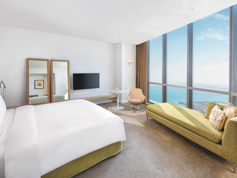 Habitación de hotel luminosa con cama grande, sofá y ventanas de piso a techo con vistas al mar.