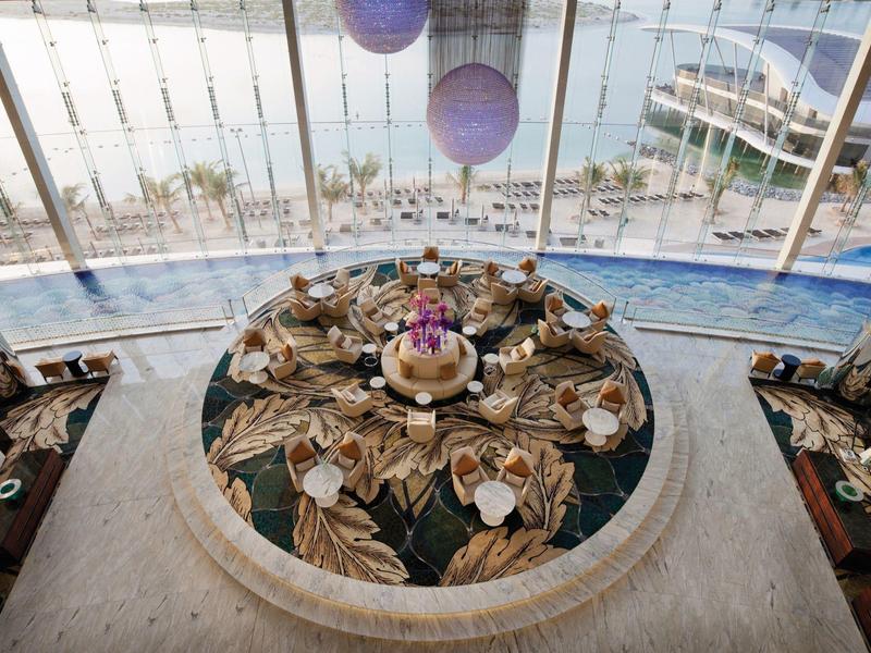 Mesa redonda con decoración floral y platos, junto a ventanales grandes con vista al mar.