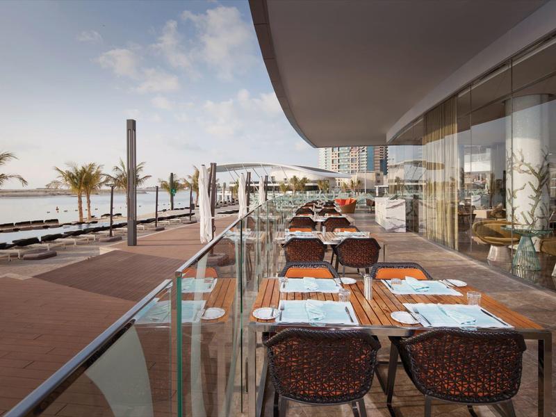 Terraza de hotel junto al mar con mesas y sillas para huéspedes, con moderna barandilla de vidrio.