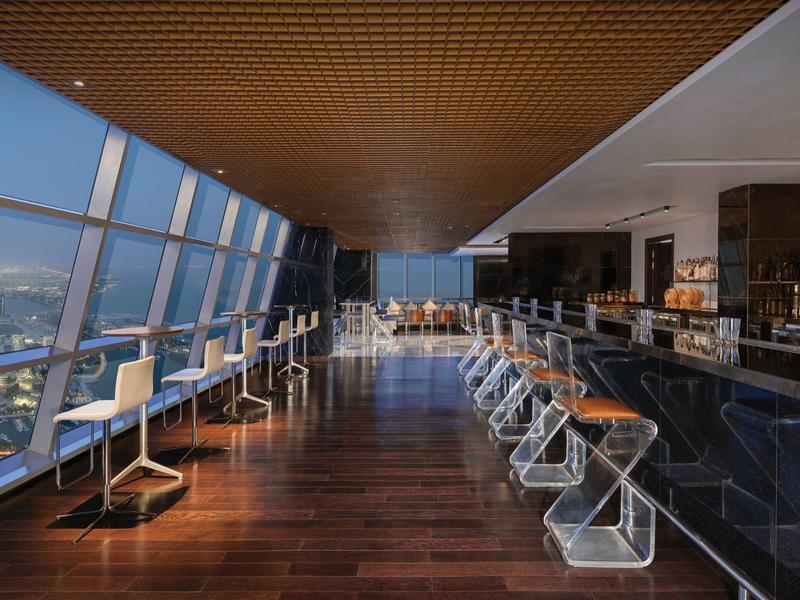 Área moderna de bar de hotel con ventanas altas y vista a la ciudad durante el día.