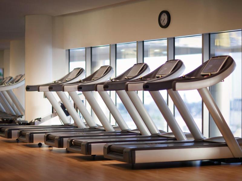 Una fila de cintas de correr en un gimnasio moderno con grandes ventanas y luz natural.