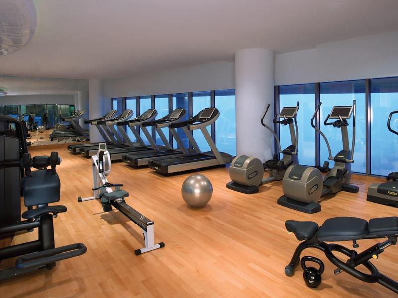 Gimnasio moderno con suelo de madera, equipos de ejercicio y ventanas panorámicas con vista exterior.