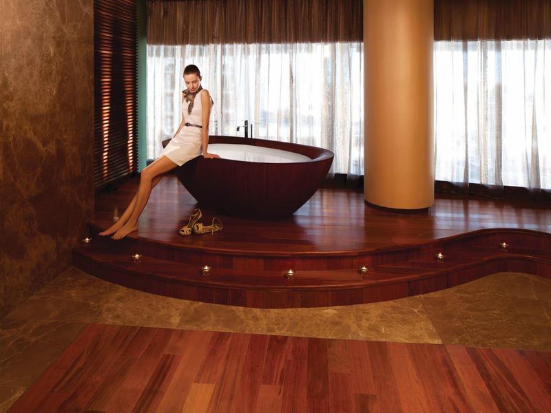 Mujer sentada en una bañera marrón sobre un podio elevado frente a cortinas del suelo al techo en un entorno hotelero.
