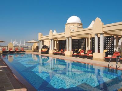 Moderní hotelový bazén s lehátky a slunečníky pod jasnou modrou oblohou.