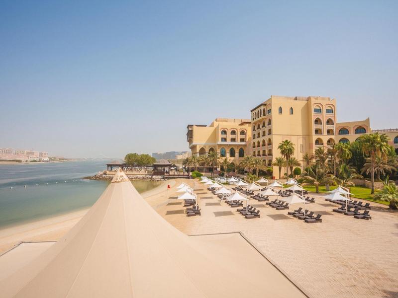 Vista de un hotel frente a la playa con sillas de playa y una zona de sombra bajo un cielo despejado.