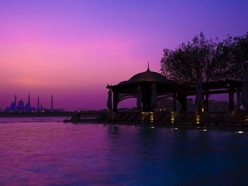 Vista nocturna de un bar de playa iluminado con piscina y cielo púrpura-naranja.