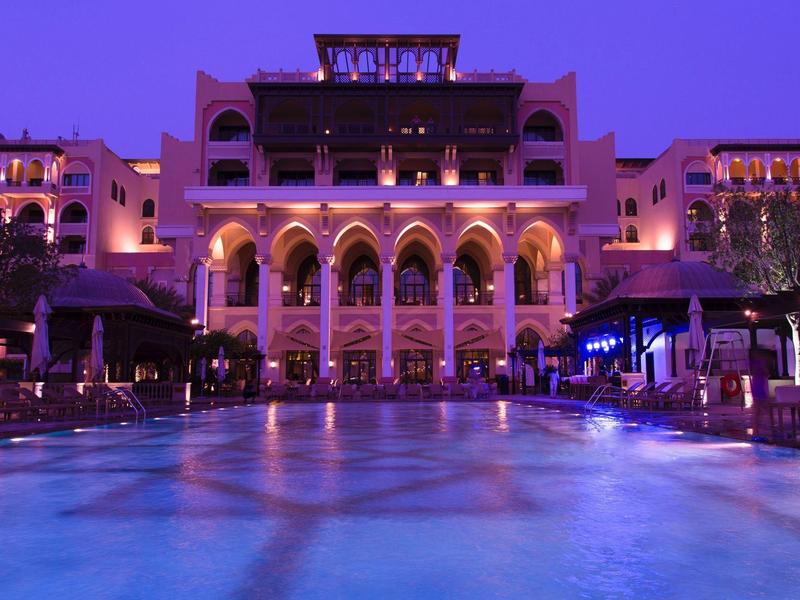 Vista nocturna de un hotel iluminado con una gran piscina en primer plano.