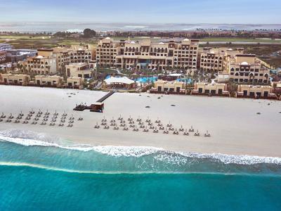 Strand mit türkisfarbenem Meer, Liegestühlen und großem Hotel mit Pool im Hintergrund.