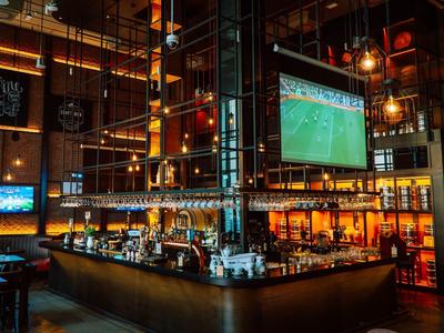 Große Bar in moderner Halle mit Fußballspiel auf Bildschirmen und warmem Lichtambiente.