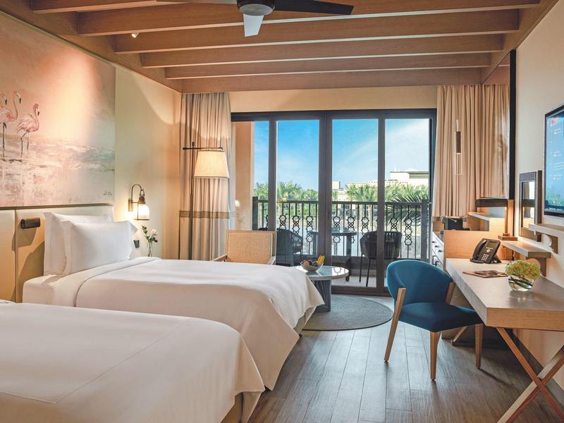 Modern hotel room with two single beds, balcony, and view of greenery.