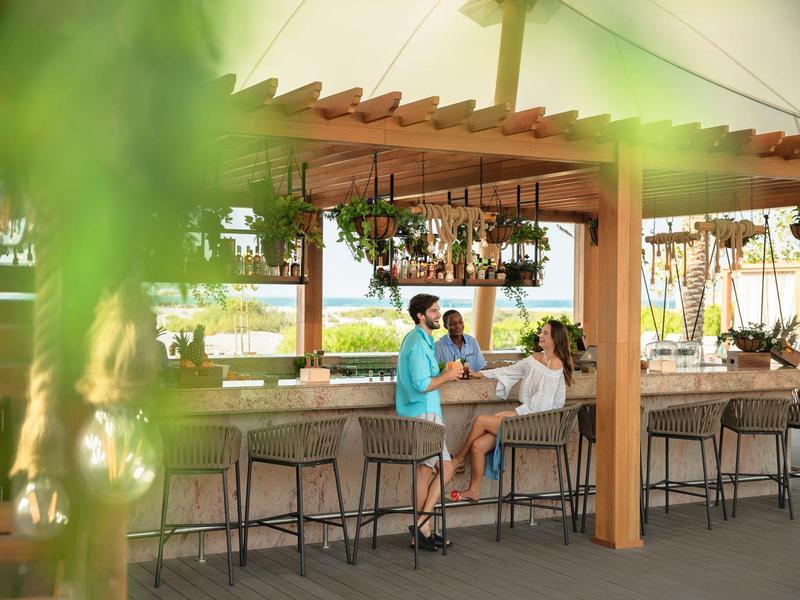 Three people sit at an outdoor bar with water views and hanging plants.