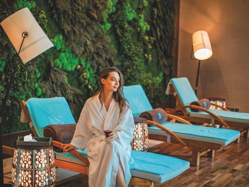 Woman in bathrobe sits on lounge chair in front of moss wall in a relaxing spa setting.