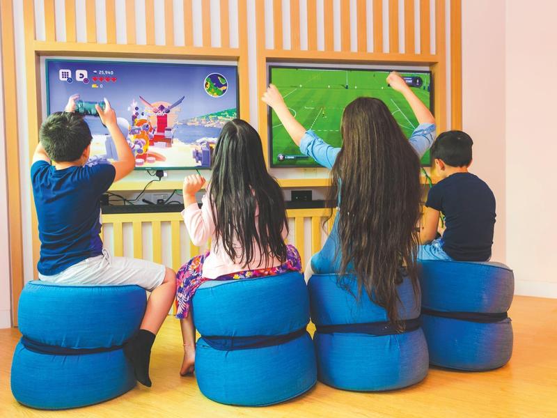 Four children sit on blue cushions playing on two large touchscreen displays.