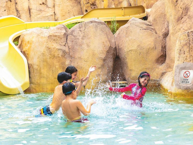 Children play and splash in the pool near a yellow water slide and rocks.