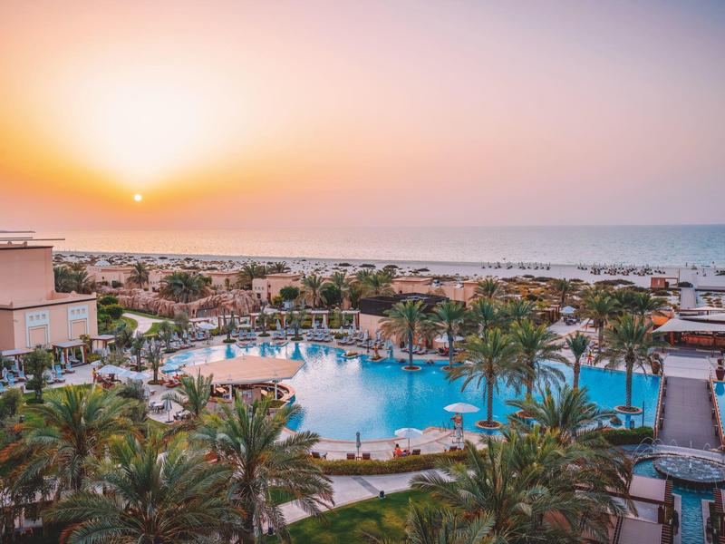View of a sunset over a resort with pool and palm trees by the sea.