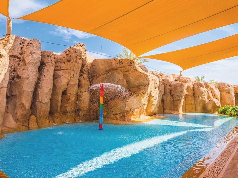 Children's pool with water play under orange sunshades and rocky landscape.