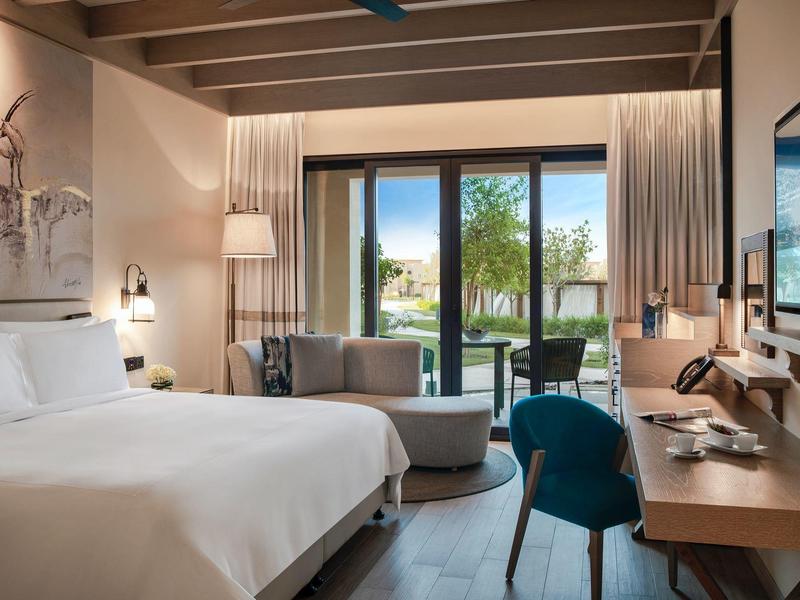 Modern hotel room with double bed, desk, and balcony overlooking a garden.