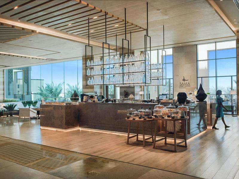 Modern hotel restaurant with large counter, snack trolley, and natural light through large windows.