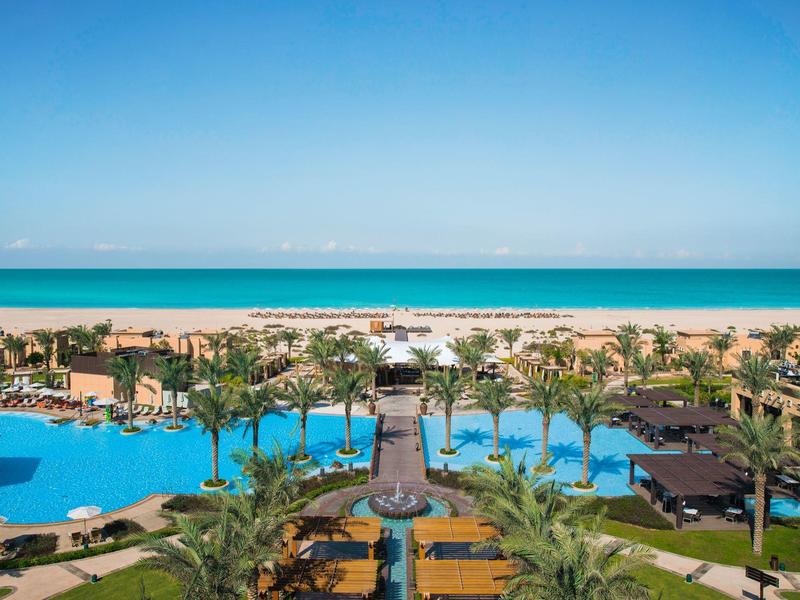 View of a tropical hotel pool with palm trees and beach under clear blue sky.