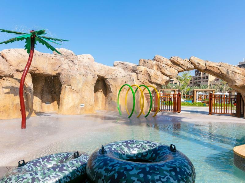 Water park with artificial palm tree, rubber rings, and rocky backdrop under sunny sky.