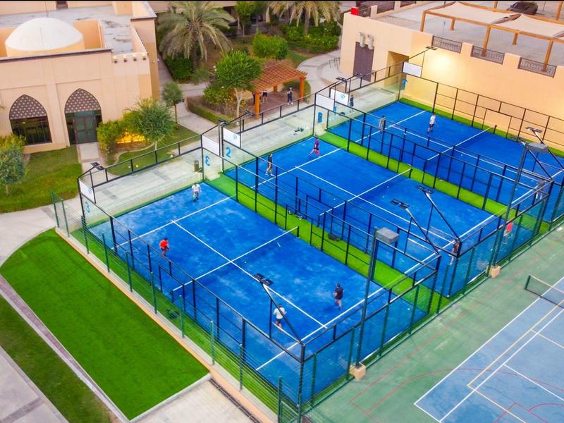 Three blue padel tennis courts at a hotel with green surroundings and nearby buildings.