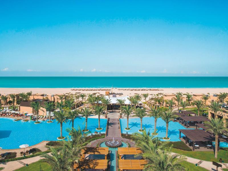 Large pool with palm trees in front of a sandy beach and blue sea sky