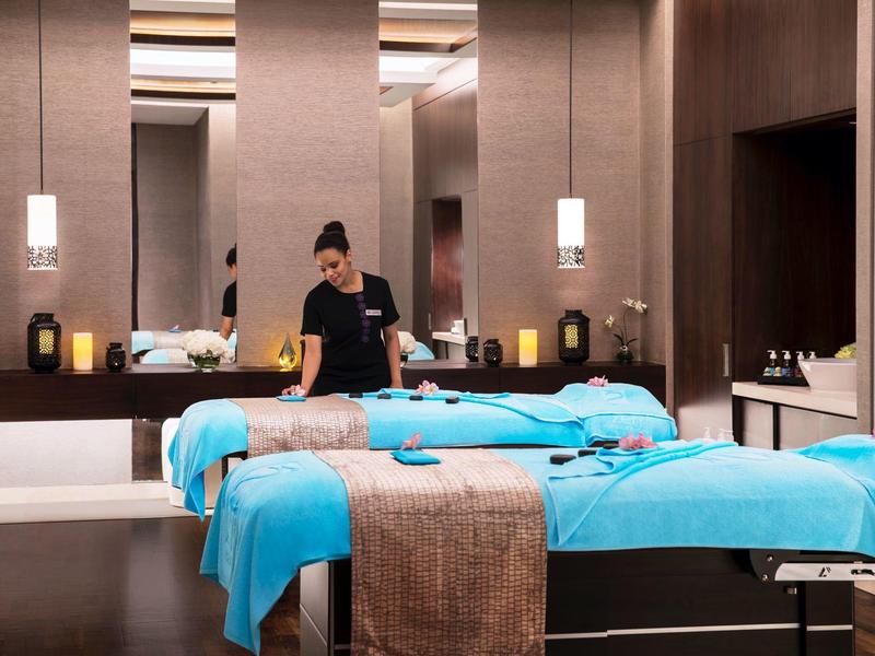 A spa room with two massage tables covered in blue towels and a woman preparing one for treatment.