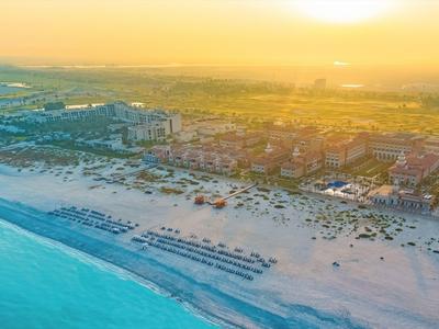 Strand mit blauem Meer und Sonnenuntergang, daneben Gebäude und Liegen am Sandstrand.