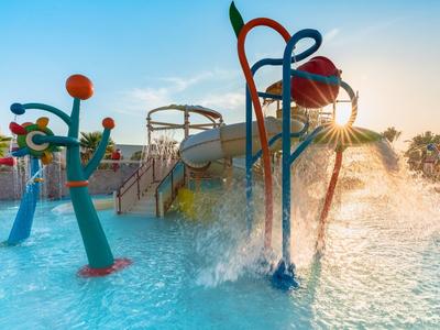 Bunter Wasserspielplatz mit Rutschen und Spritzanlagen in flachem Pool bei Sonnenuntergang.