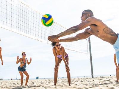 Sechs Personen spielen Volleyball am Strand, eine Person taucht ausgedehnt zum Ball.