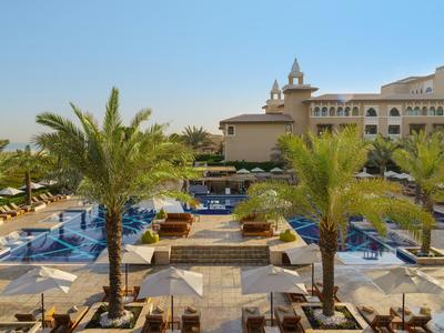 Großer Hotelpool mit Palmen, Sonnenschirmen und Liegen vor einem cremefarbenen Gebäude bei Sonnenschein.