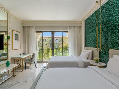 Helles Hotelzimmer mit zwei weißen Betten, grün gemusteter Wand und Balkon mit Gartenaussicht.