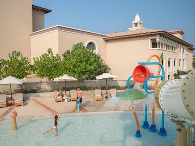 Children play in a shallow pool with water slides next to a hotel building.
