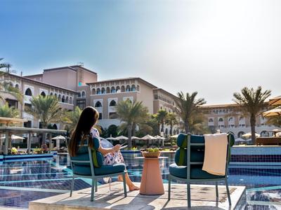 Woman relaxes by the pool with palm trees and a large hotel building in sunlight.