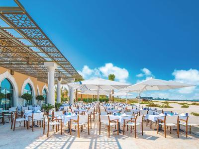 Terrassenrestaurant mit weißen Tischen, Stühlen, Sonnenschirmen und Blick auf das blaue Meer.