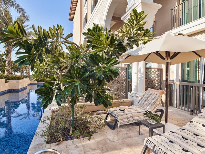 Outdoor lounge chairs and umbrella beside a pool at a sunny hotel patio with tropical plants.