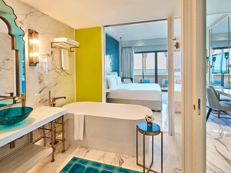 Modern hotel suite with bathtub, glass basin, and bedroom with large windows and balcony.
