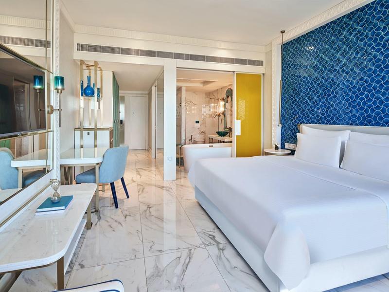 Modern hotel room with king bed, marble floor, blue accent wall, and workspace with chair.