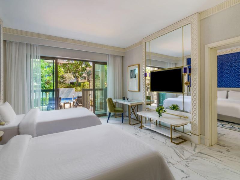 Bright hotel room with two beds, a balcony, desk, and TV, decorated in white and blue tones.