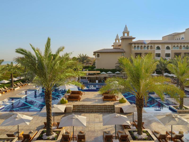 Luxury hotel pool area with palm trees, sun loungers, and blue-tiled pools under clear sky.